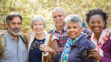 Active senior adult friends hiking in wooded forest area. The multi-ethnic group wears a backpack and uses binoculars to explore nature around them. Middle Eastern, Latin, African and Caucasian ethnicities. 60s to 70s age range.
