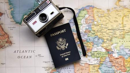 A travel-themed flat lay image featuring a vintage Kodak Instamatic camera, a United States passport, and a world map. The passport is placed in the center, slightly overlapping the camera, with the camera's strap draped across the map. The map showcases parts of Europe, North Africa, and the Middle East, emphasizing a sense of adventure and global exploration.